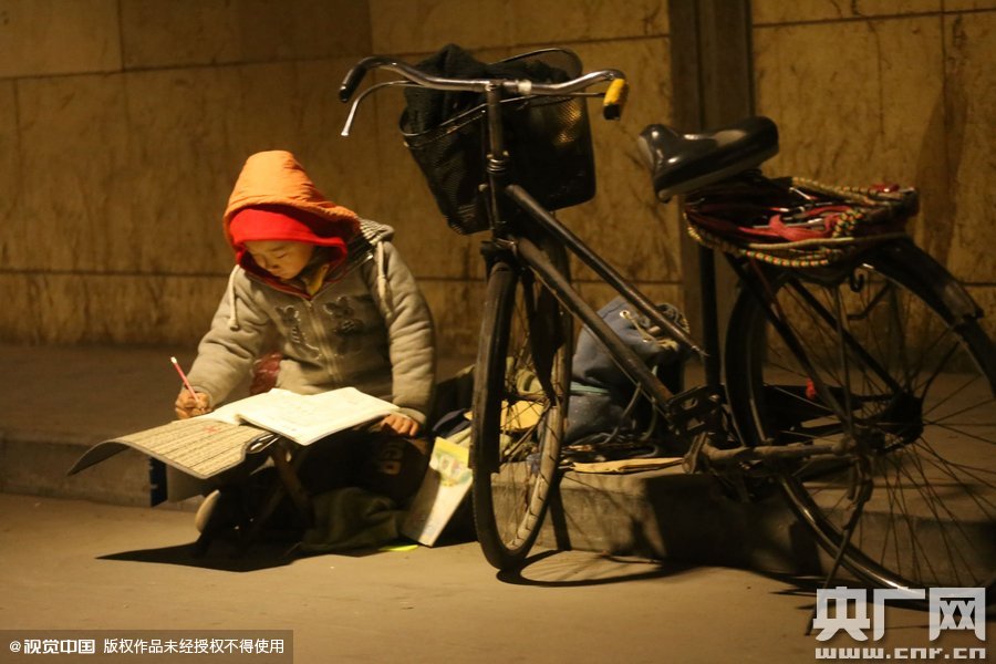 鄭州中原路京廣鐵路大橋下涵洞的馬路牙子邊，一位戴著紅色辣椒帽的小男孩趴在一個小馬扎上，上面鋪著一張車墊，在微弱的燈光下寫作業(yè)